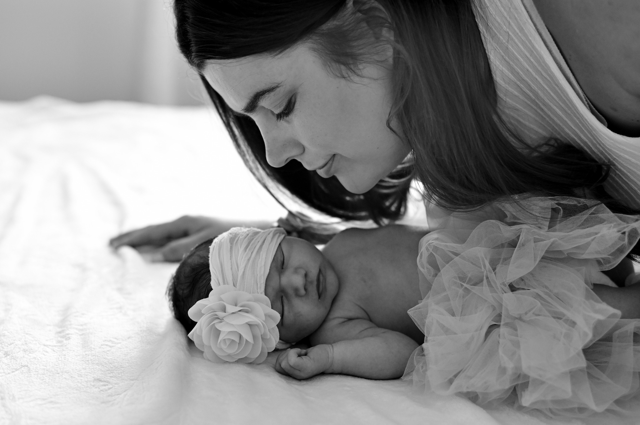 Czarno-białe zbliżenie: matka czule spogląda na śpiące noworodka z kwiatem na opasce, delikatne światło podkreśla intymność momentu.