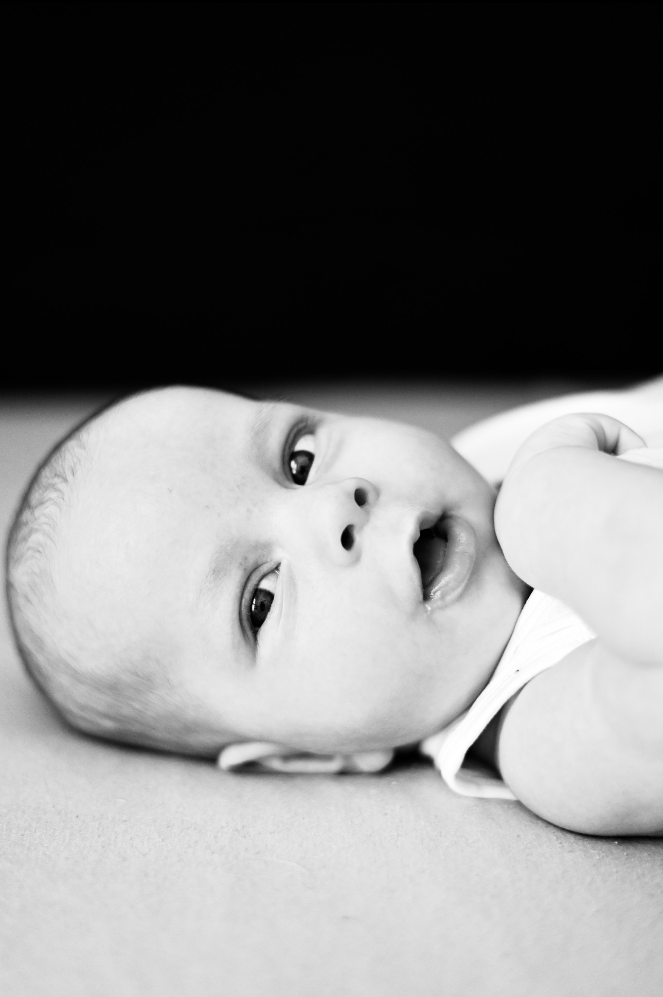 Czarno-białe zbliżenie na twarz leżącego noworodka z otwartymi ustami i dużymi oczami, skierowanymi w górę, na jasnym tle.
