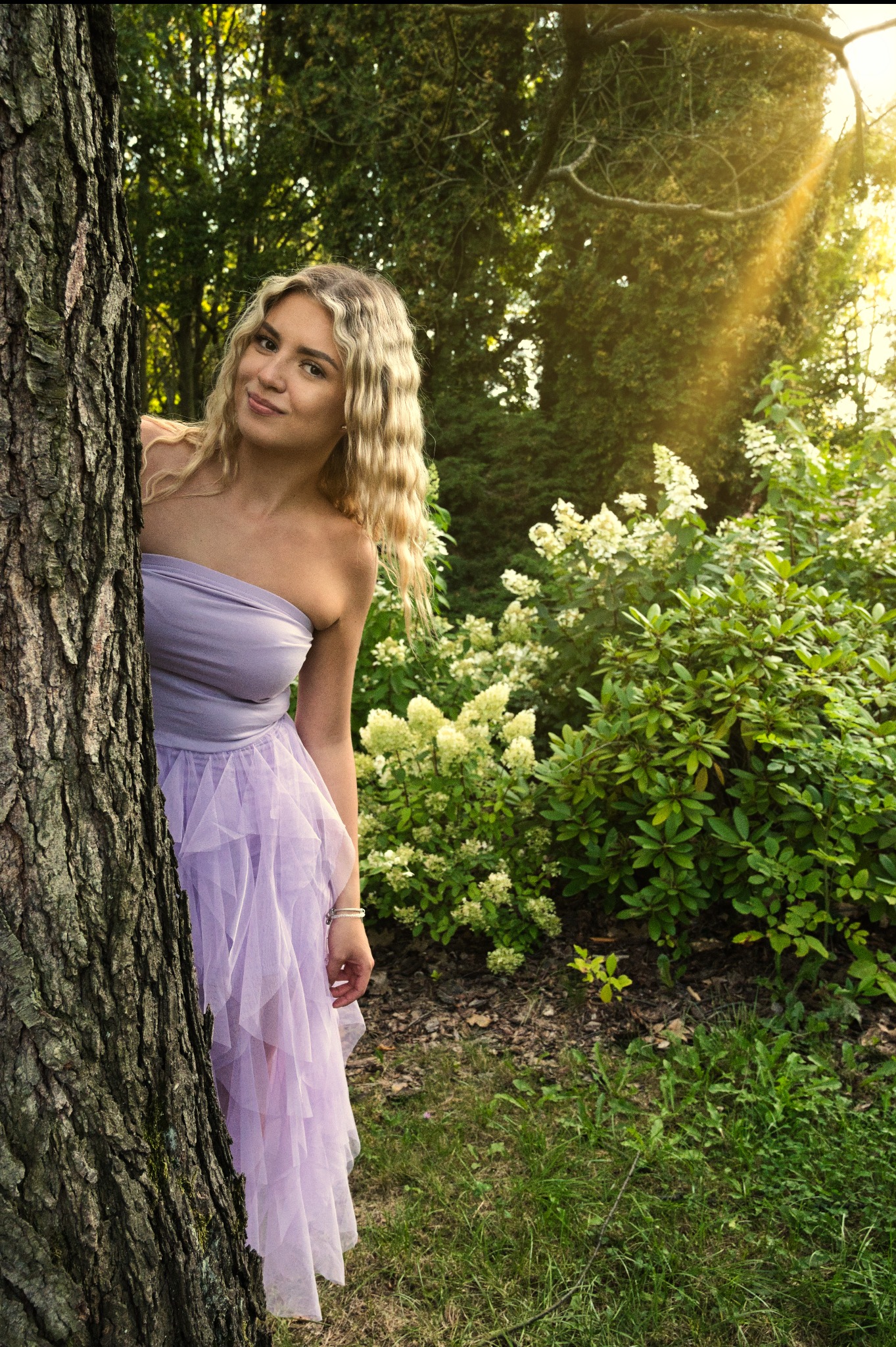 Uśmiechnięta blondynka w liliowej sukience, częściowo schowana za drzewem w słonecznym ogrodzie. Romantyczna sceneria, naturalne światło.