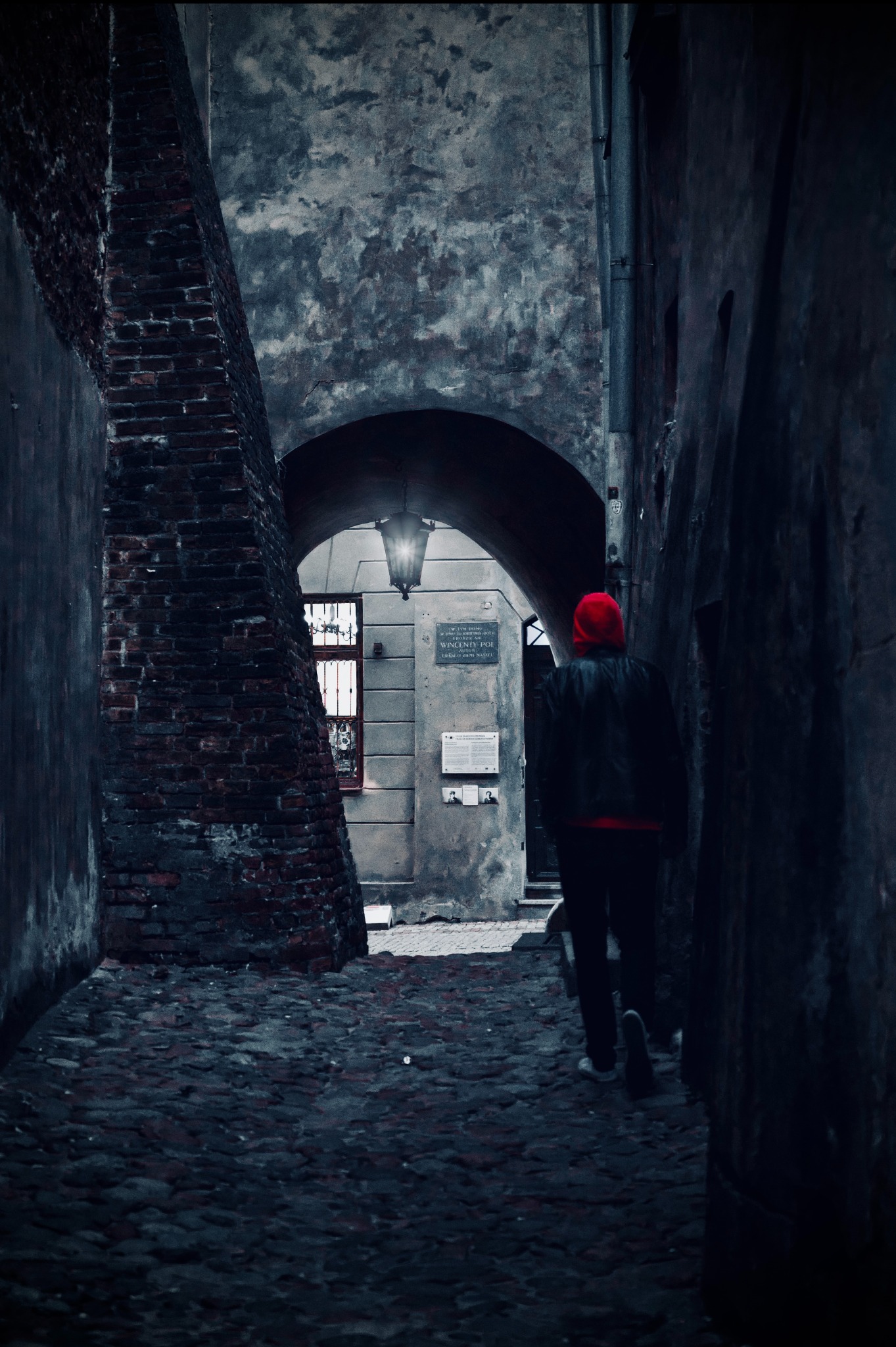 Mroczne ujęcie wąskiej uliczki z brukowaną nawierzchnią, gdzie postać w czerwonej czapce oddala się w kierunku łuku z oświetleniem i tablicą pamiątkową.