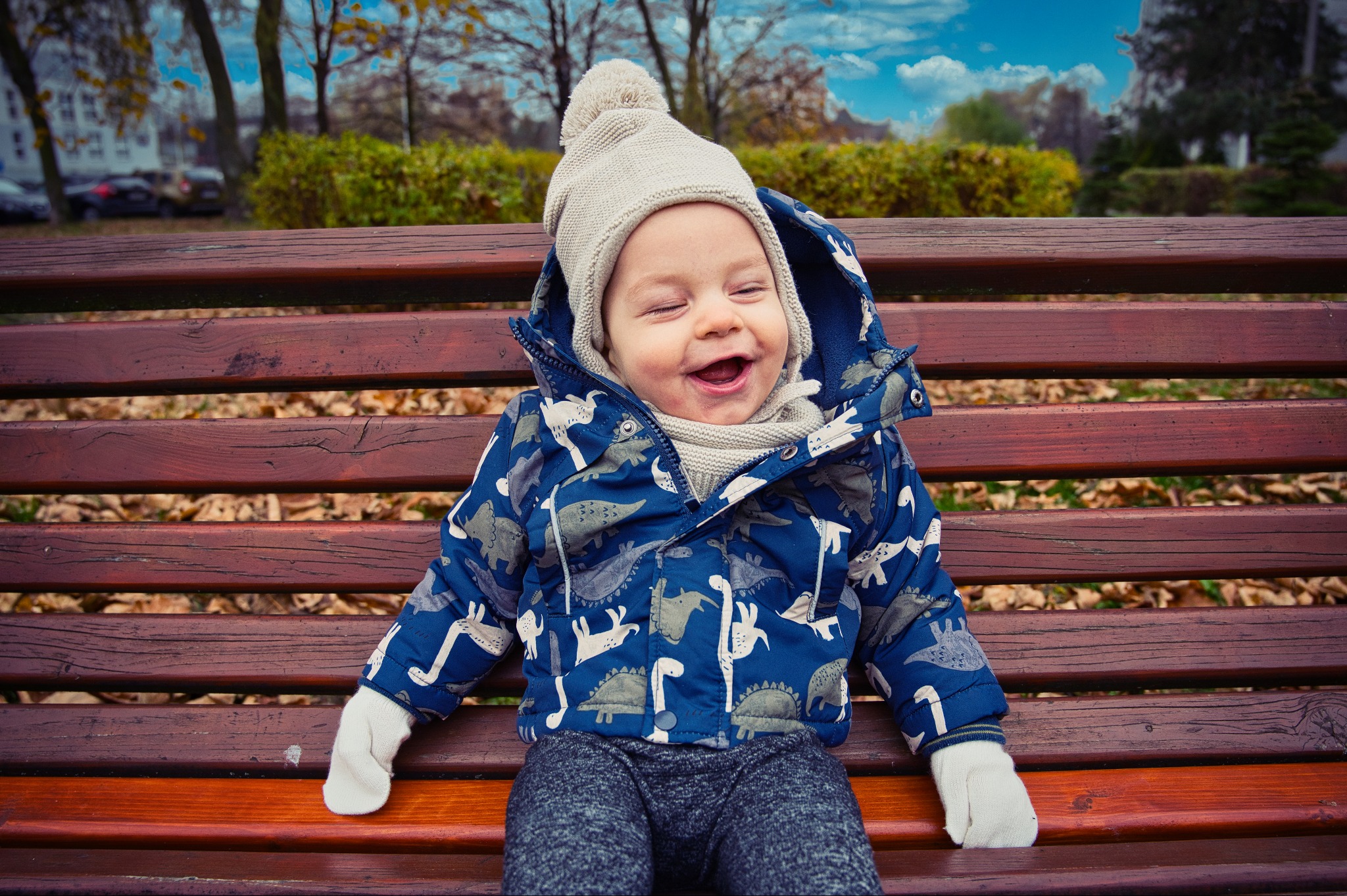 Uśmiechnięte dziecko w zimowej kurtce i czapce siedzi na drewnianej ławce w parku jesienią. W tle drzewa i błękitne niebo z chmurami.