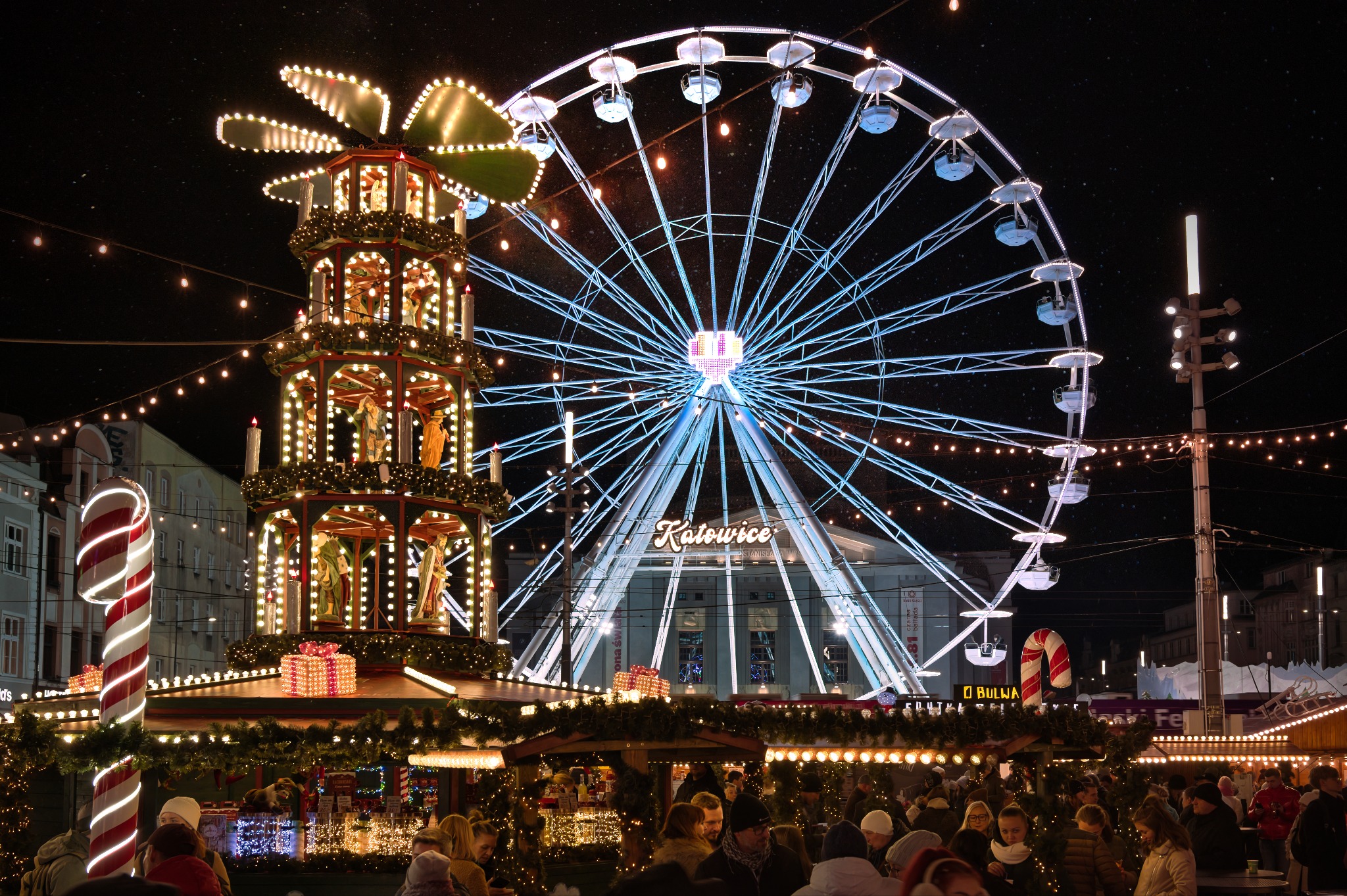 Nocny widok na jarmark bożonarodzeniowy w Katowicach: oświetlony młyn adwentowy, diabelski młyn z napisem 'Katowice' i tłum ludzi w dole.