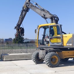 AEM TRANS Mariusz Maj - Żółta koparka kołowa Volvo EW140B podnosi betonowe płyty na placu budowy. Widoczne stalowe liny i chwytak. W tle ogrodzenie i zieleń.