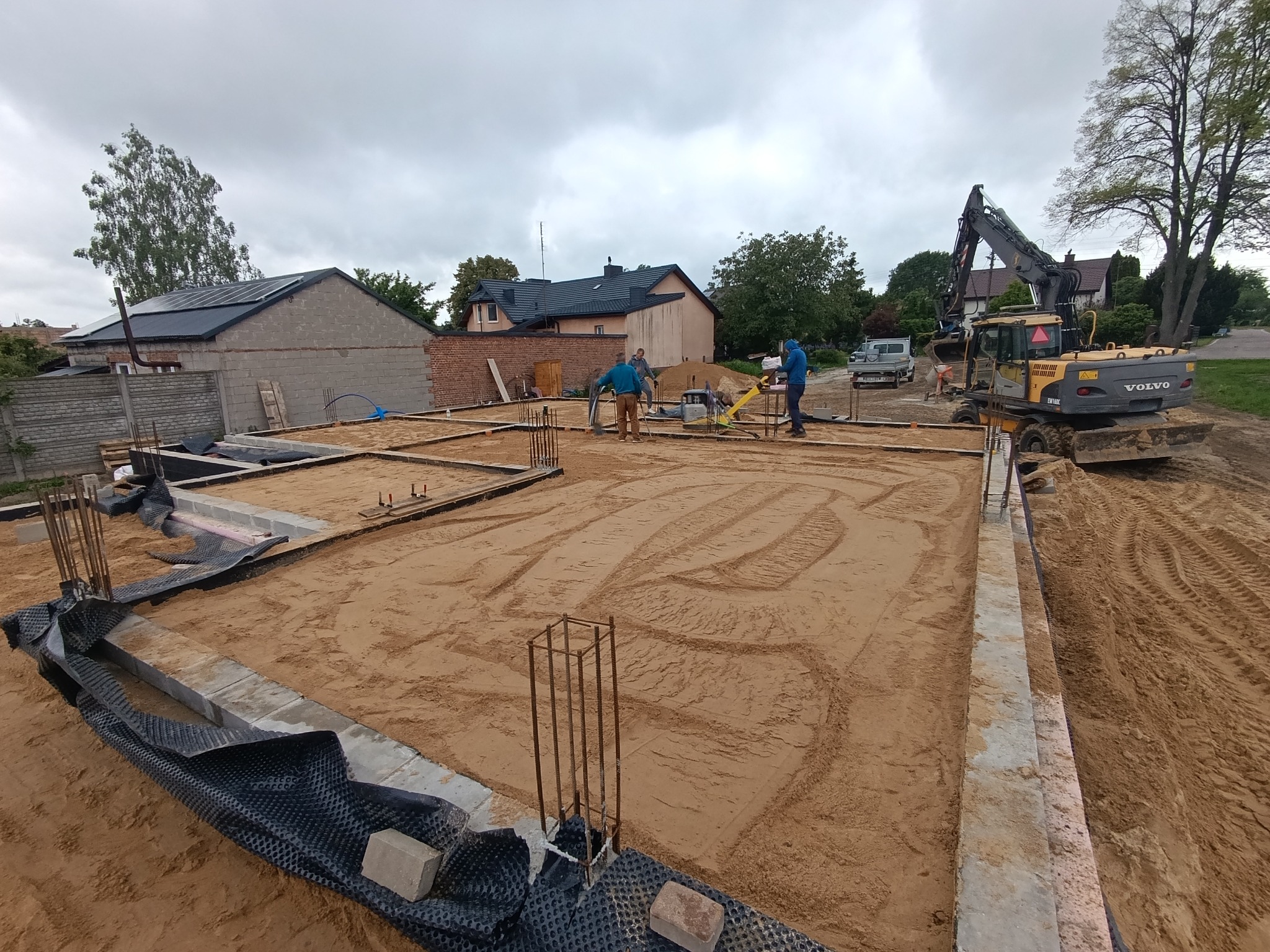 Przygotowanie terenu pod fundamenty: wyrównany piasek, szalunki, zbrojenia, ekipa budowlana i koparka Volvo na działce z domami w tle.