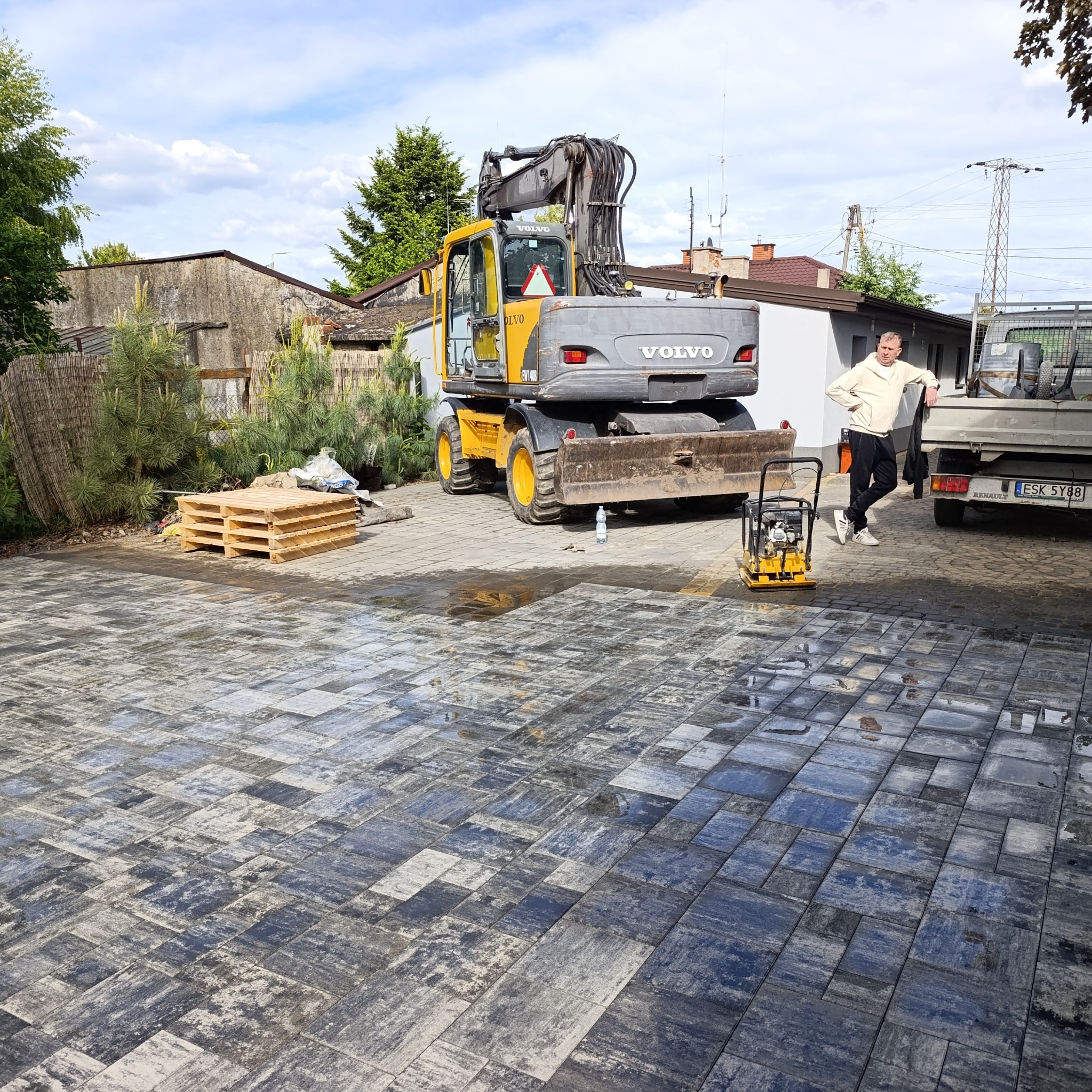 Ułożona kostka brukowa w szaro-niebieskich odcieniach. W tle żółta koparka Volvo, ubijarka i mężczyzna oparty o samochód dostawczy. Paleta z kostką obok.