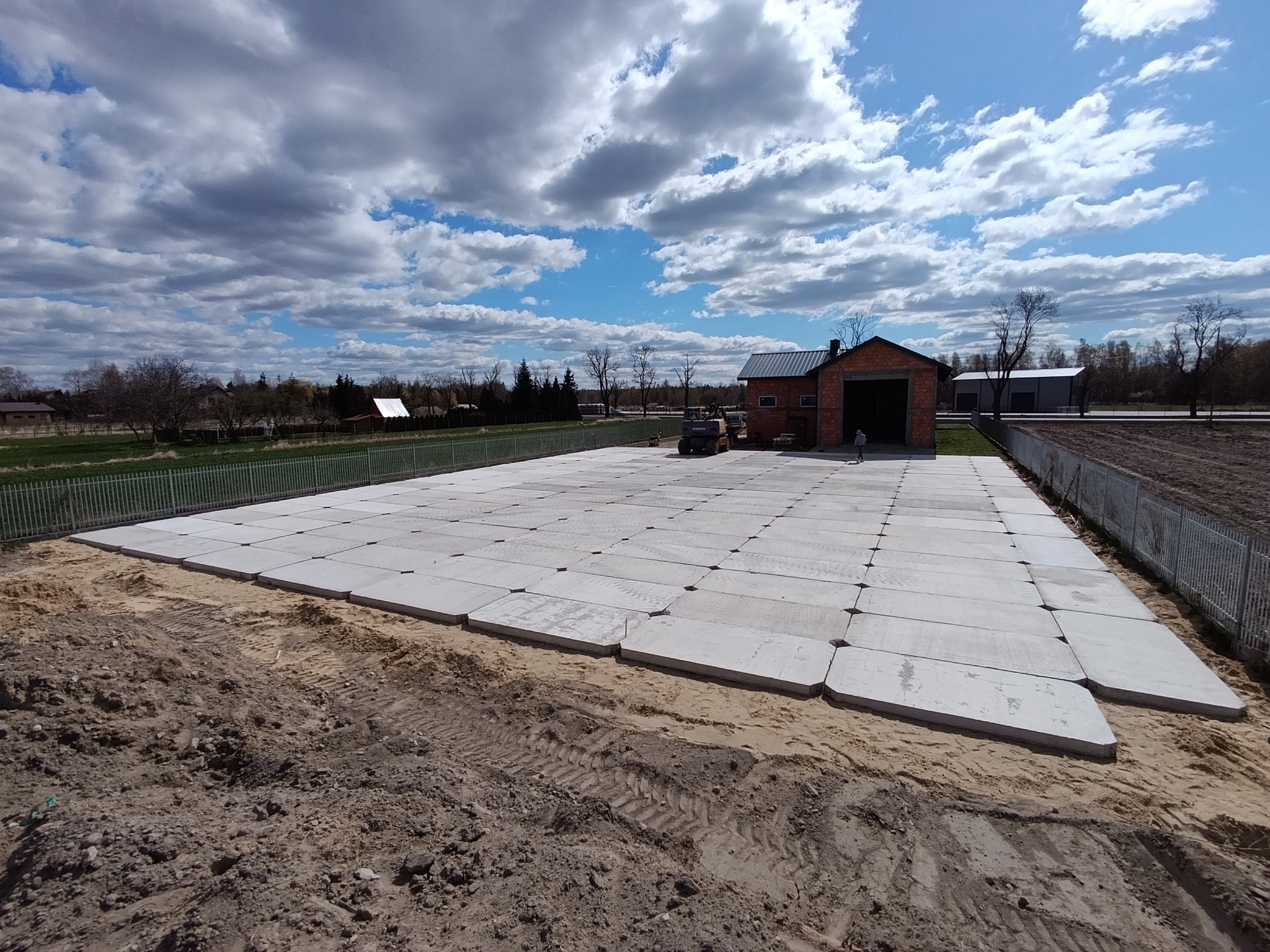 Rozległy plac wyłożony betonowymi płytami, z widocznym budynkiem w tle i sprzętem budowlanym. Całość otoczona ogrodzeniem, na tle błękitnego nieba z chmurami.