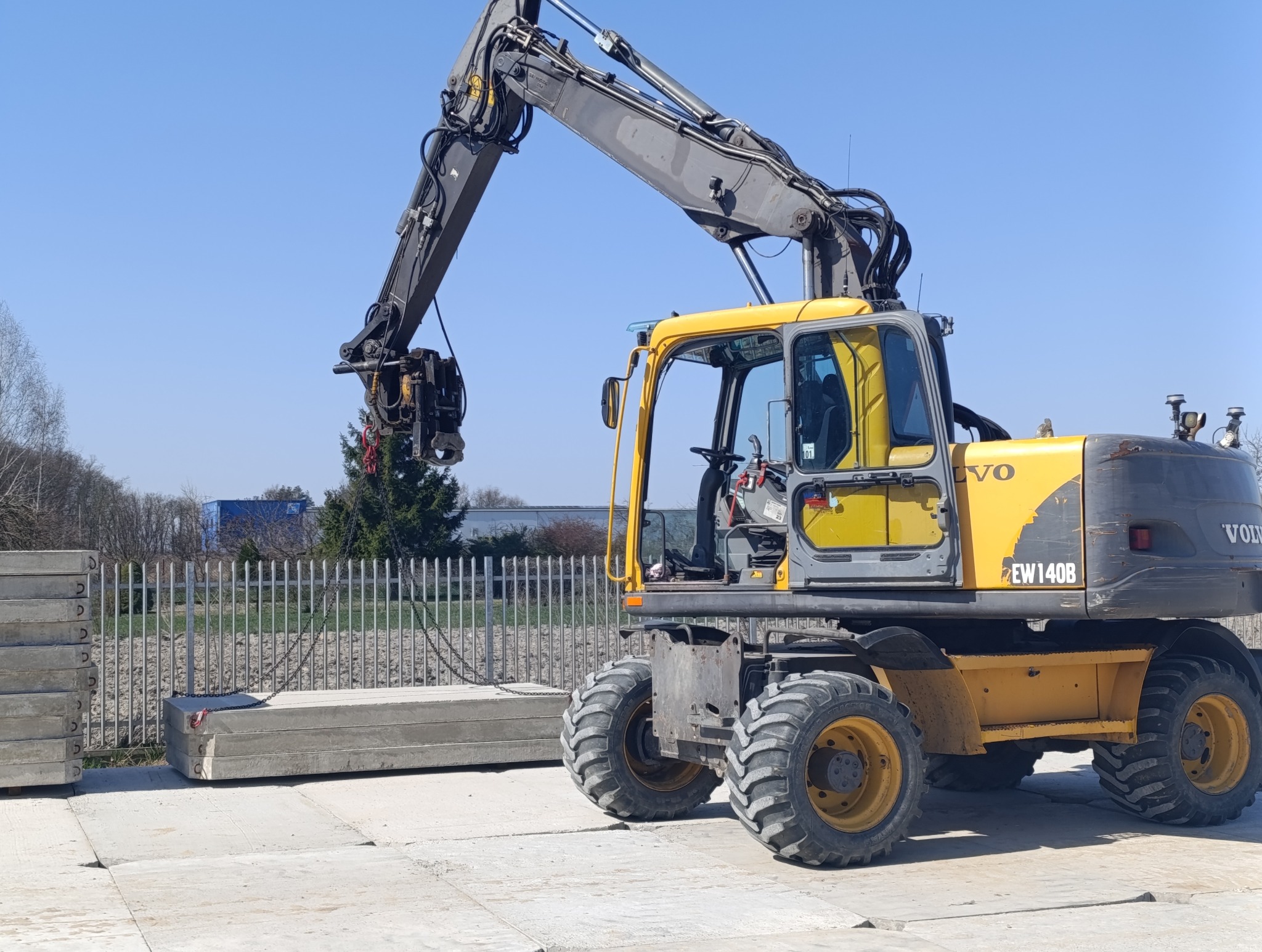 Żółta koparka kołowa Volvo EW140B podnosi betonowe płyty na placu budowy. Widoczne stalowe liny i chwytak. W tle ogrodzenie i zieleń.