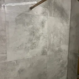 Ściana kabiny prysznicowej wyłożona szarymi, matowymi płytkami imitującymi beton, z widoczną złotą armaturą prysznicową i delikatnym odbiciem światła na powierzchni płytek.