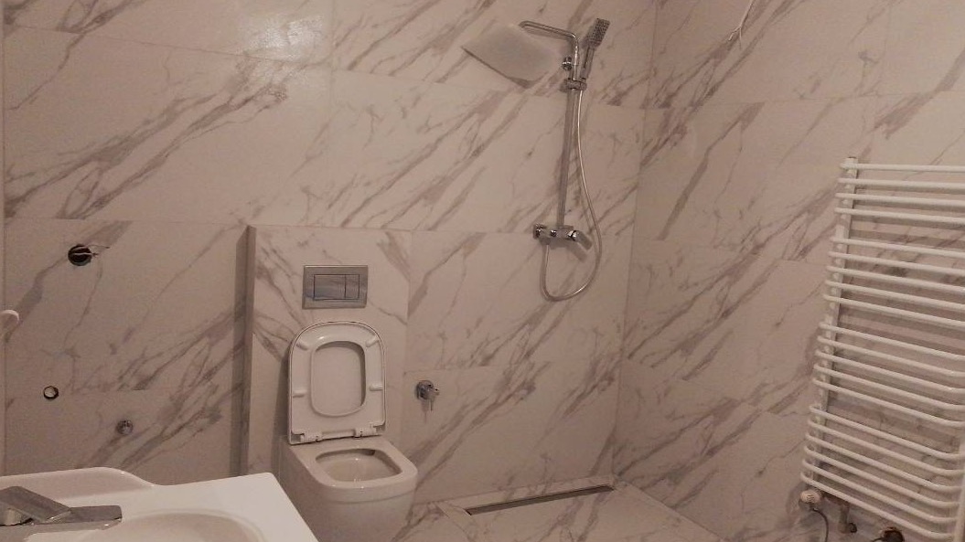 Wykończona łazienka z marmurowymi płytkami, wiszącą toaletą z otwartą klapą i grzejnikiem drabinkowym.