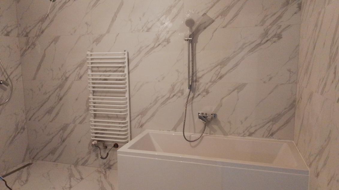 Wykończona łazienka z białą wanną, grzejnikiem drabinkowym i marmurowymi płytkami na ścianach i podłodze, widok z boku.