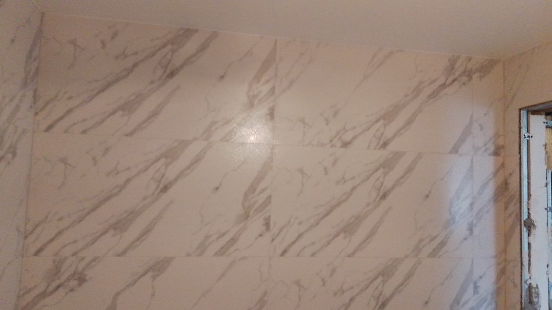 Ściana wyłożona białymi płytkami imitującymi marmur, widoczne fugi i fragment metalowej ramy drzwi.