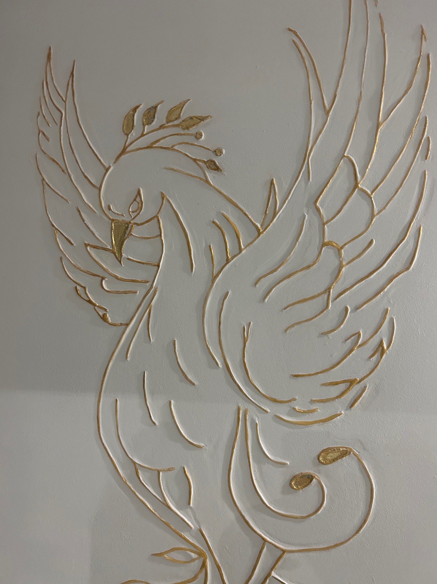 Dekoracyjna płaskorzeźba przedstawiająca feniksa z gałązką oliwną na białej ścianie, wykonana ze złotej masy szpachlowej.