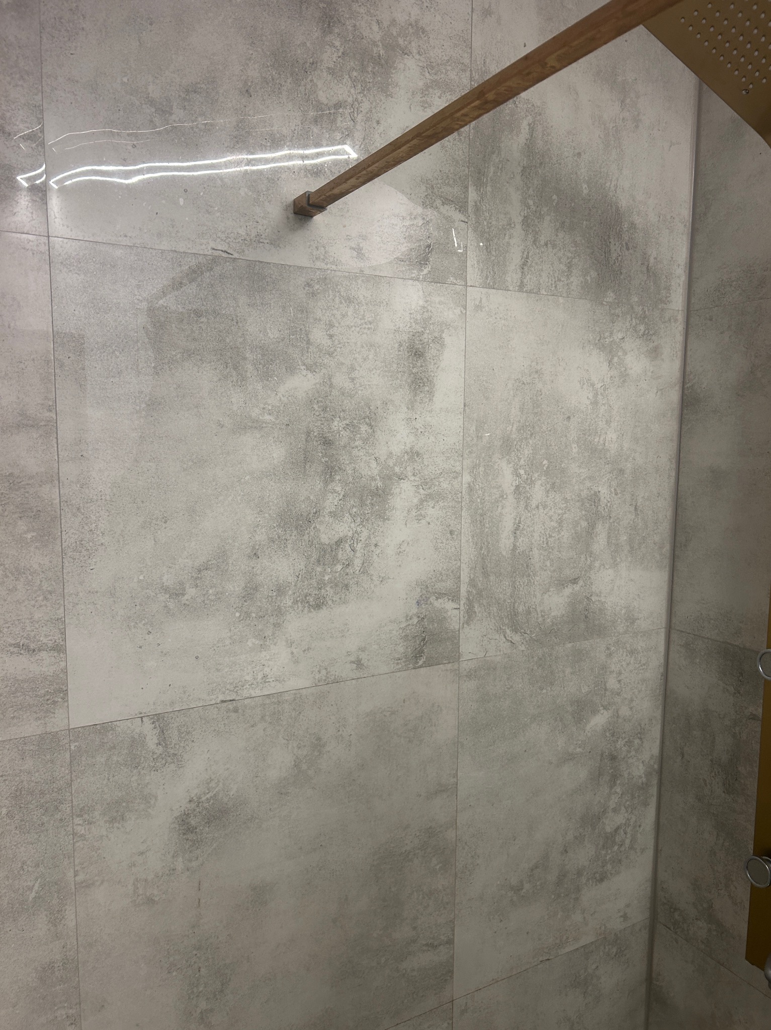 Ściana kabiny prysznicowej wyłożona szarymi, matowymi płytkami imitującymi beton, z widoczną złotą armaturą prysznicową i delikatnym odbiciem światła na powierzchni płytek.