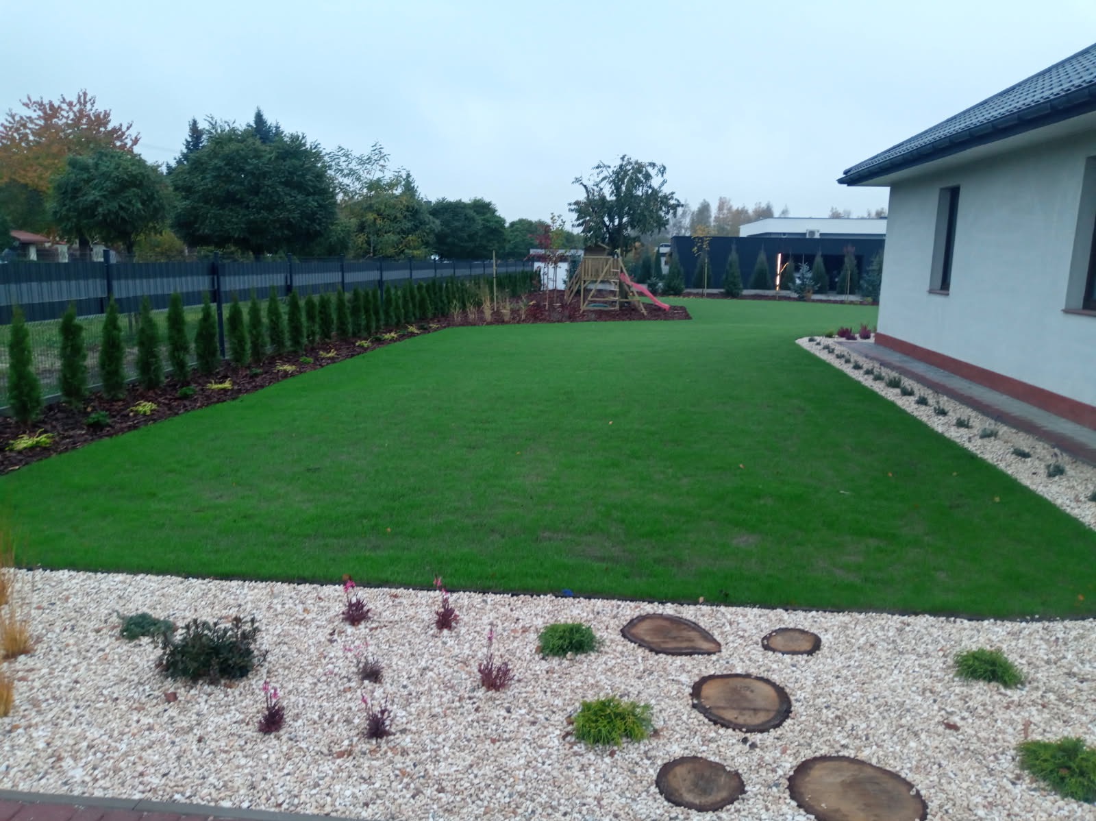 Zadbany ogród z równo przystrzyżonym trawnikiem, ozdobnym żwirem, drewnianymi krążkami i tujami wzdłuż ogrodzenia, w tle plac zabaw z huśtawką i zjeżdżalnią.