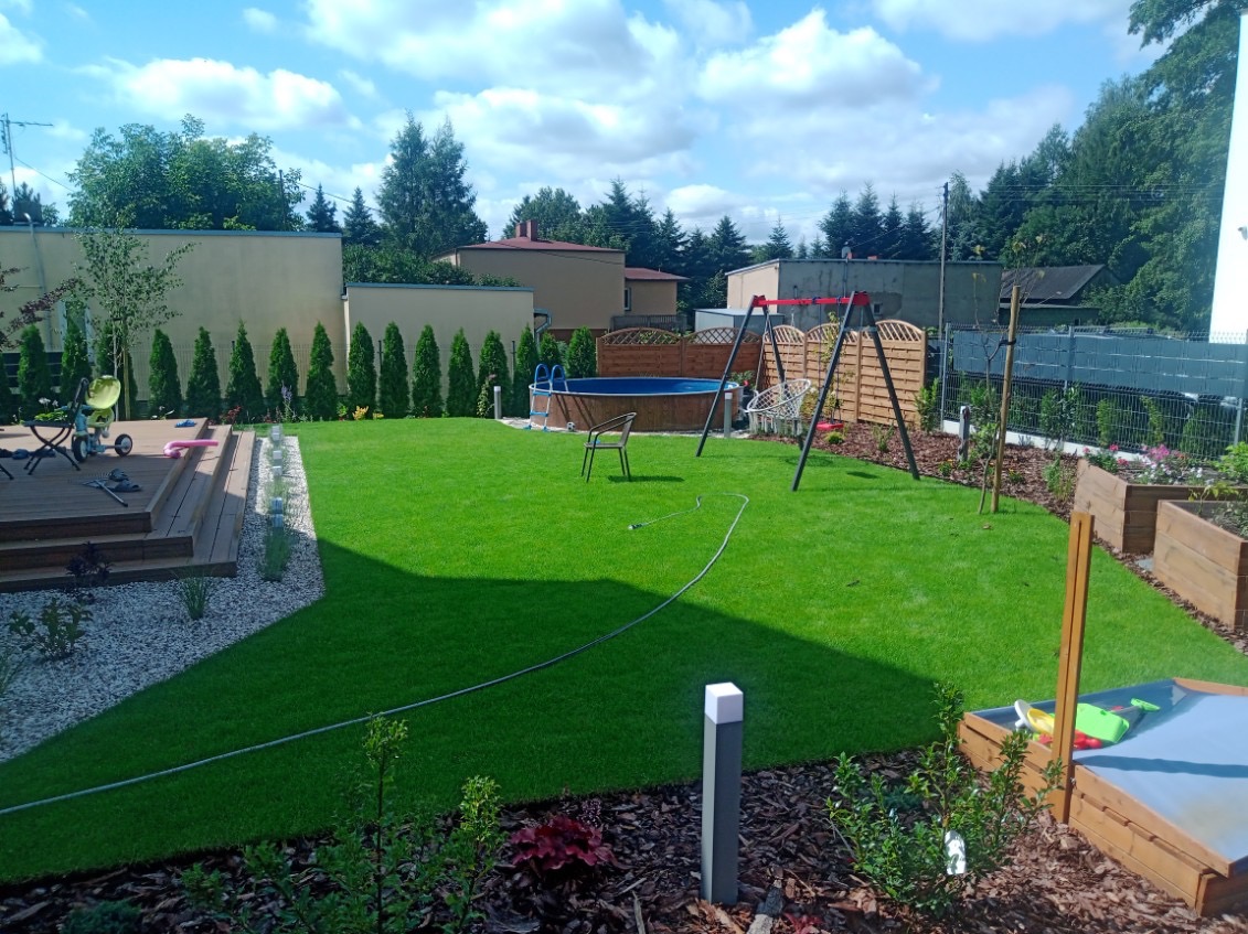 Zadbany ogród z równo przystrzyżonym trawnikiem, drewnianym tarasem, basenem, huśtawką i podwyższonymi rabatami, otoczony tujami i ogrodzeniem.