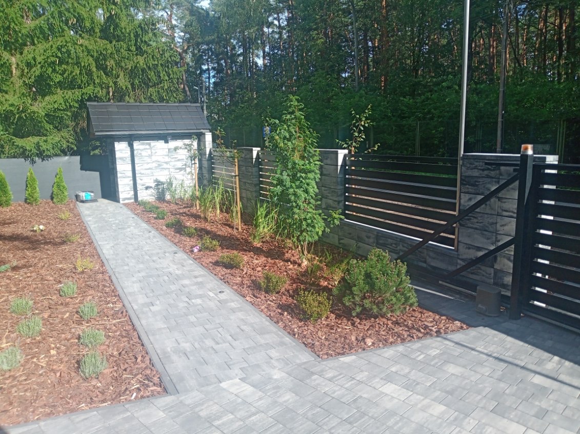 Nowoczesny ogród z szarym chodnikiem z kostki, ciemnym ogrodzeniem panelowym i świeżo posadzonymi roślinami, kora rozsypana na rabatach.