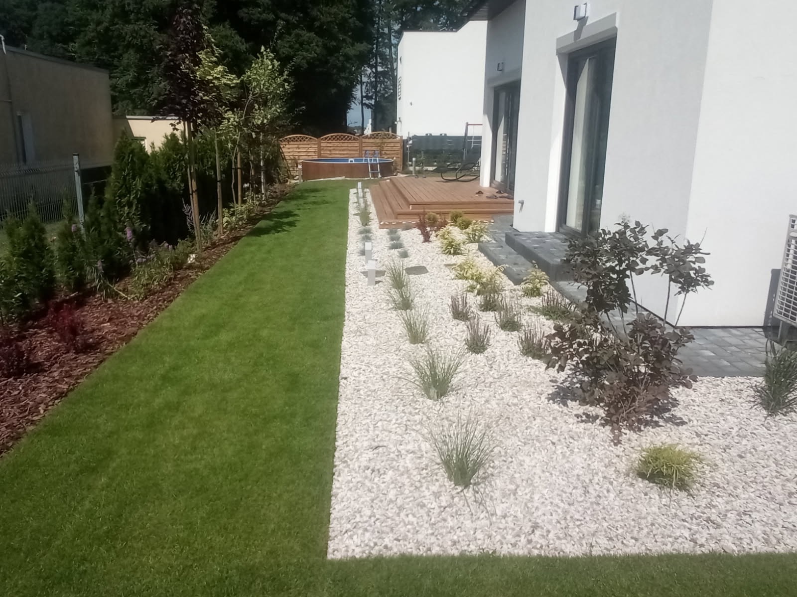 Nowoczesny ogród z białym żwirem, zielonym trawnikiem i drewnianym tarasem z basenem w tle.