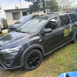 Jacek Szupryczyński - Czarny samochód typu kombi z napisem 'TAXI XL BYDGOSZCZ' na boku, stojący na trawie, z widocznym budynkiem w tle.