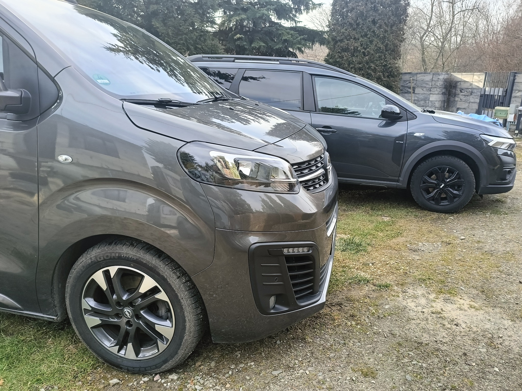 Dwa szare samochody osobowe: Citroen SpaceTourer i Dacia Duster, stojące na żwirowym podjeździe przed domem.