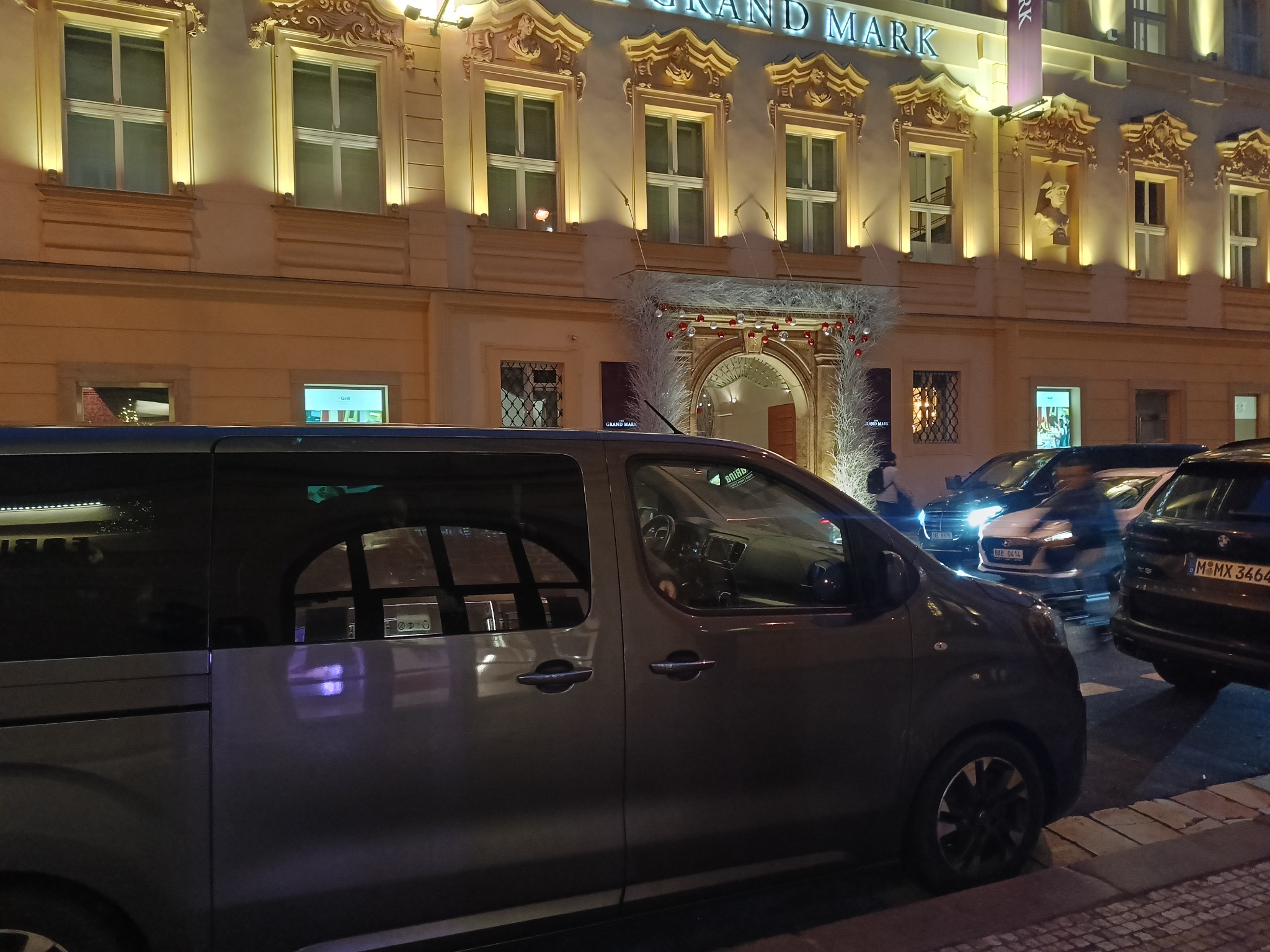 Szary bus zaparkowany przed budynkiem Grand Mark w Pradze, widoczne inne samochody w tle.