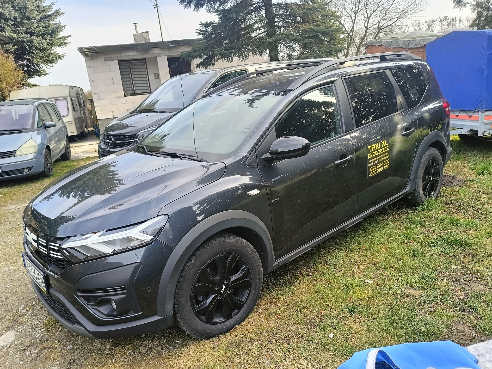 Czarny samochód typu kombi z napisem 'TAXI XL BYDGOSZCZ' na boku, stojący na trawie, z widocznym budynkiem w tle.