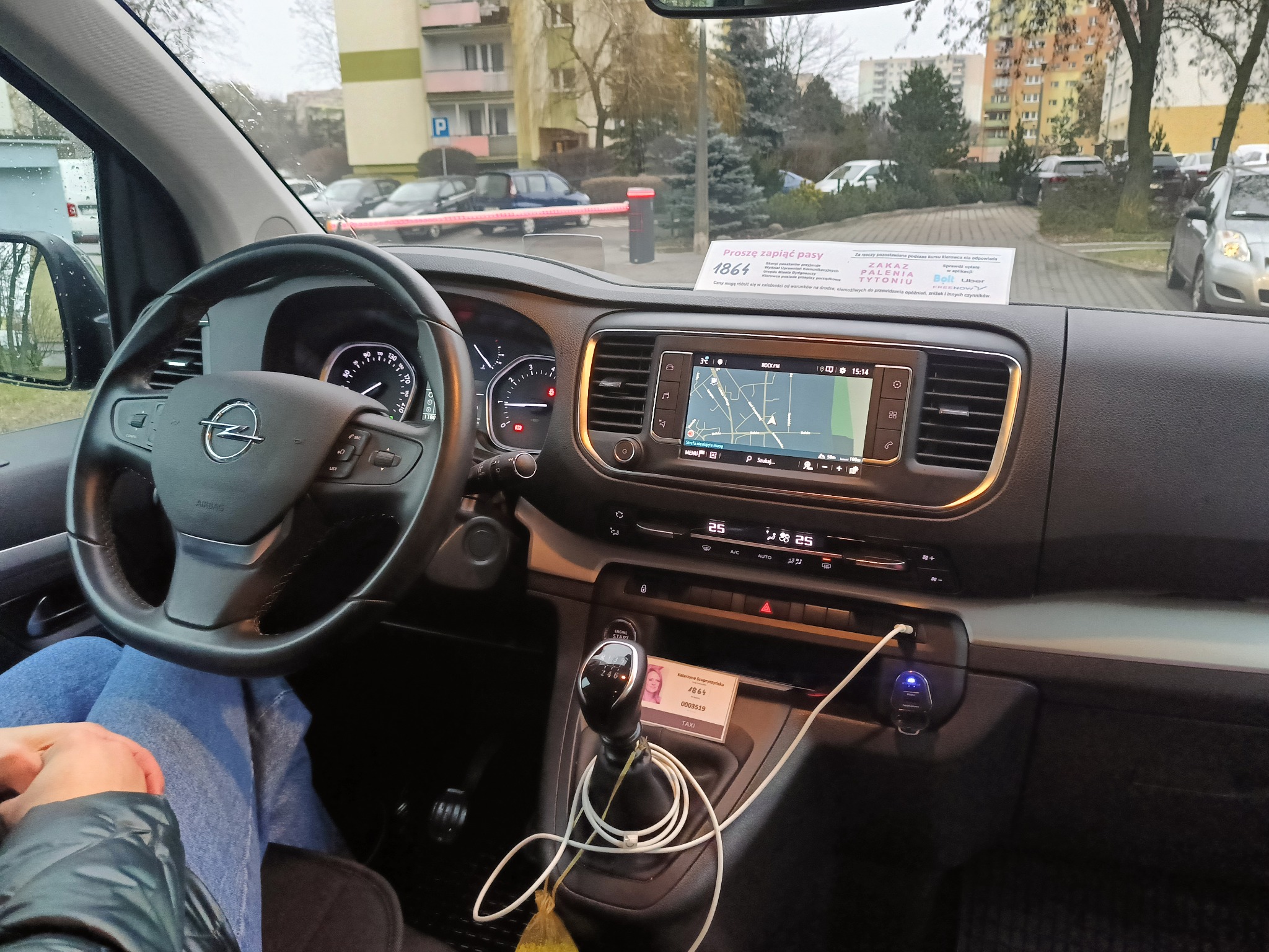 Wnętrze busa Opel z widocznym pulpitem nawigacyjnym, kierownicą z logo i kartą identyfikacyjną taksówkarza przy dźwigni zmiany biegów. Na desce rozdzielczej widoczna kartka z informacją o opłatach...