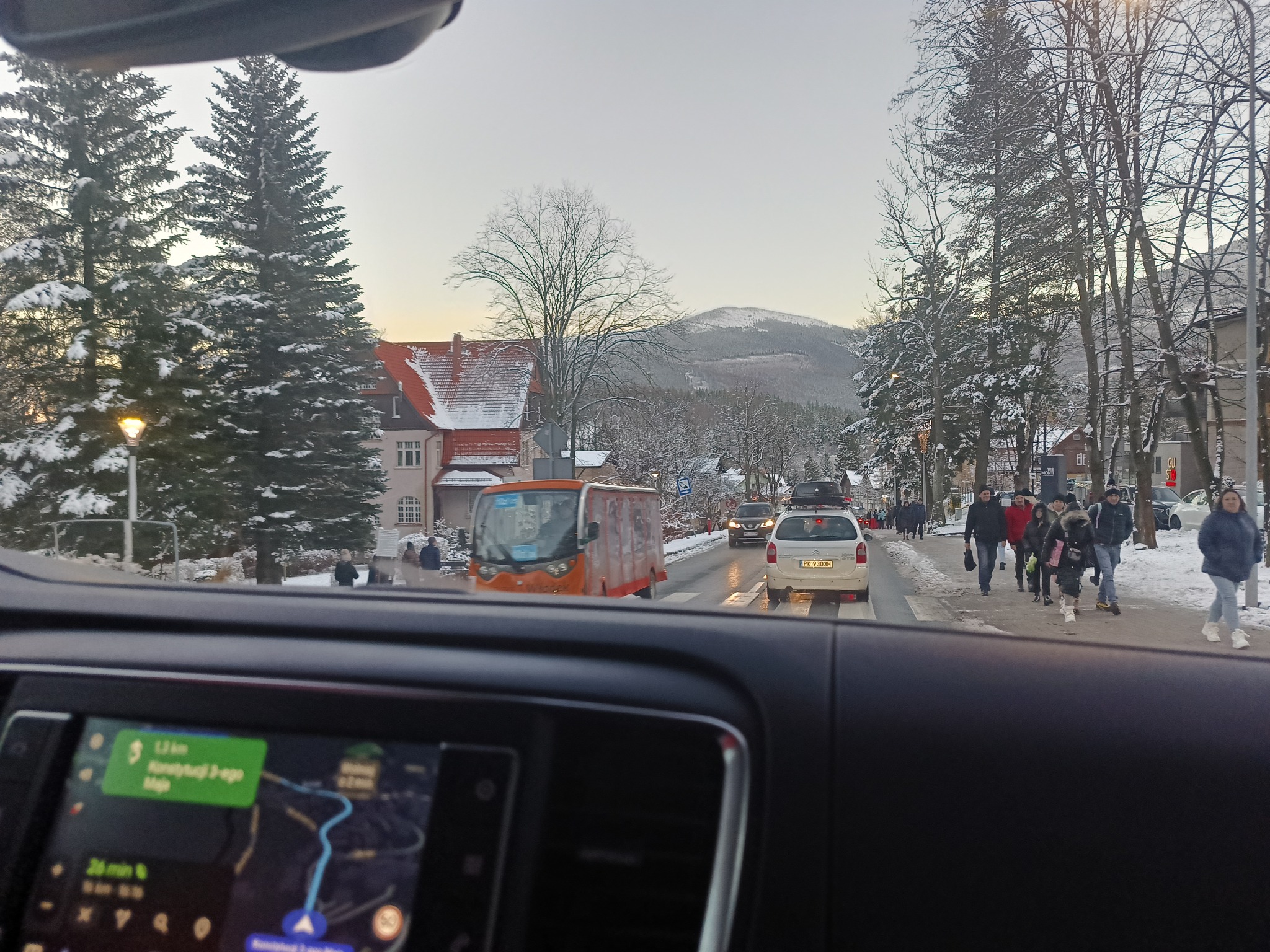 Widok z wnętrza samochodu na zaśnieżoną ulicę w górskiej miejscowości, z widocznym autobusem elektrycznym i pieszymi na chodniku, w tle góra.