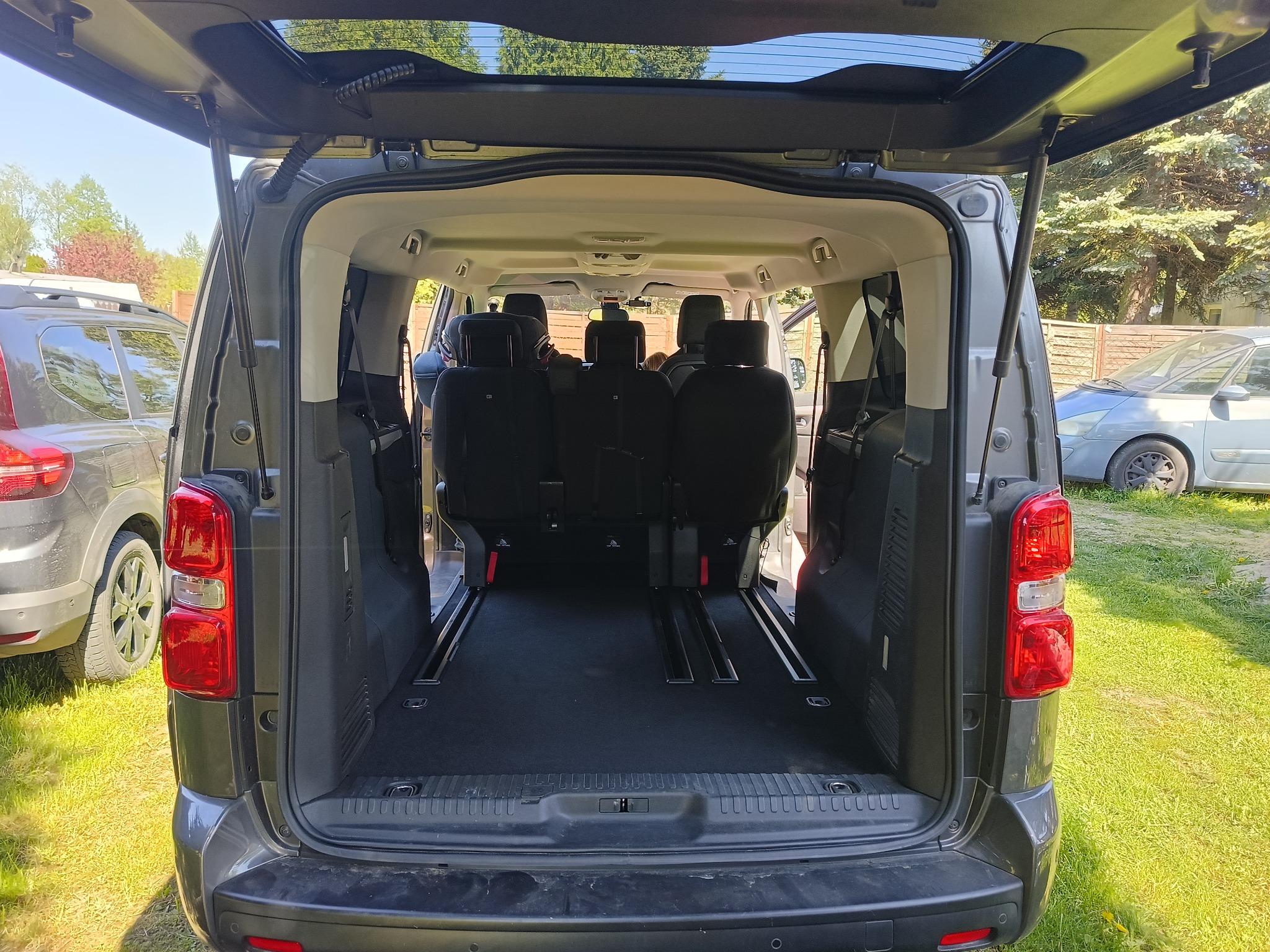 Wnętrze bagażnika szarego busa z widocznymi szynami mocującymi siedzenia, otwartą klapą i fragmentami innych pojazdów w tle, na trawniku.
