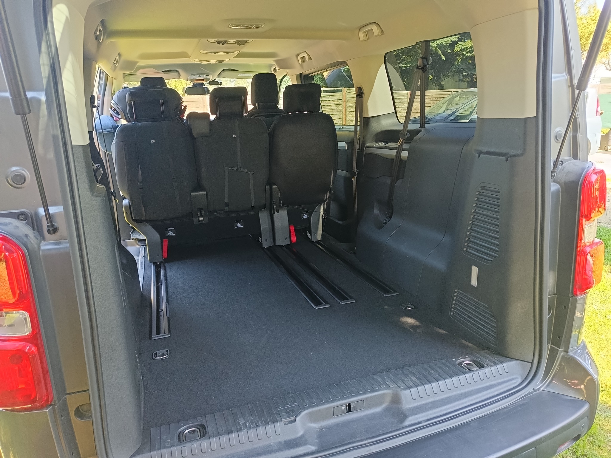 Wnętrze szarego busa z widocznymi szynami do mocowania foteli i przestrzenią bagażową. Fotele ustawione w konfiguracji dla pasażerów.