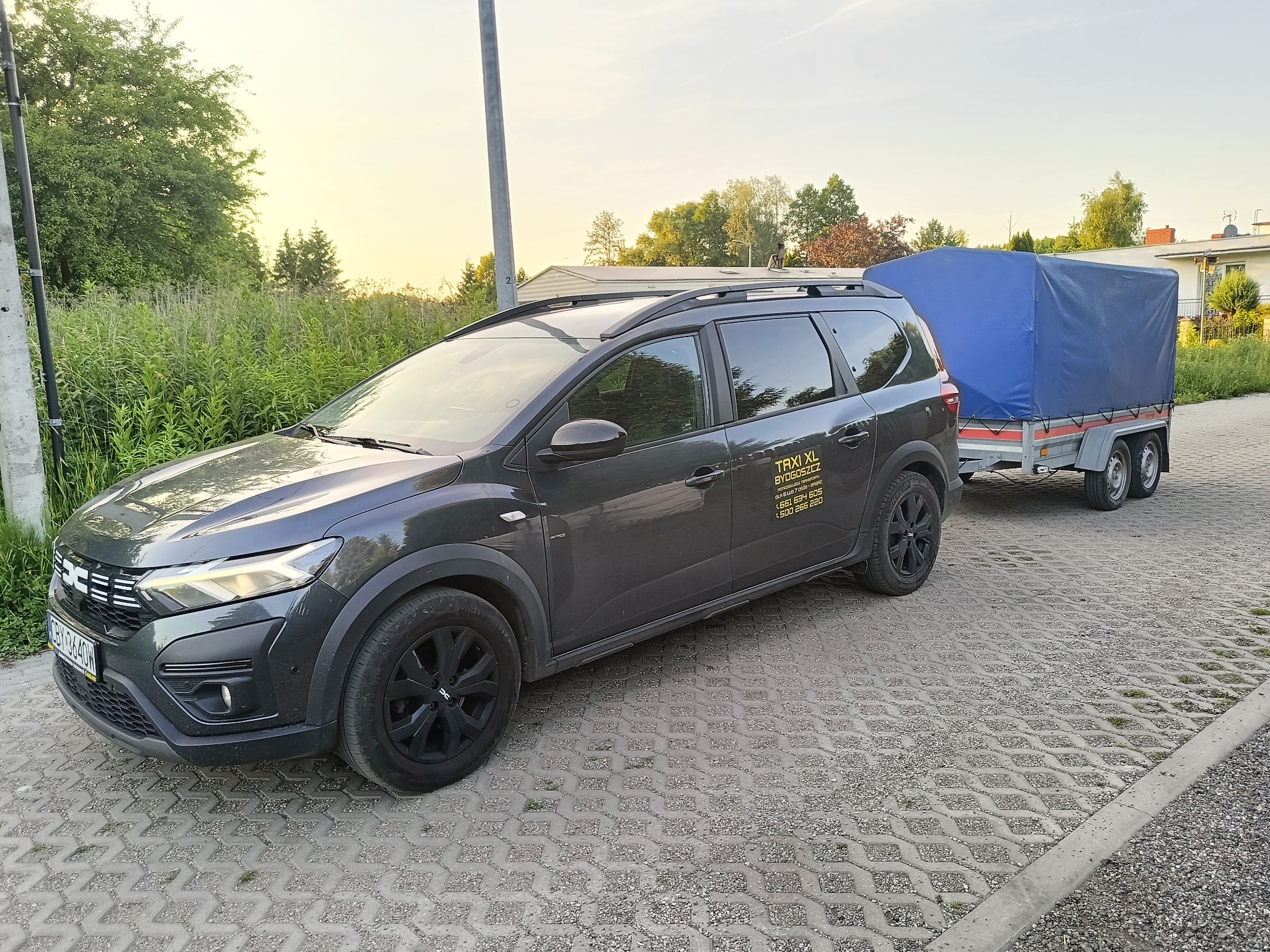 Szary samochód osobowy z przyczepą pod plandeką, Taxi XL Bydgoszcz, transport. Widok z boku na kostce brukowej, zieleń w tle, spokojne światło.