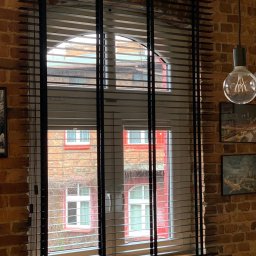 Igor Kravets - Ciemne żaluzje drewniane na tle ceglanej ściany i okna z widokiem na budynek. Lampa wisząca z żarówką Edisona po prawej stronie kadru.