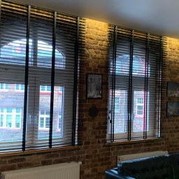 Igor Kravets - Dwa okna z czarnymi żaluzjami drewnianymi na tle ściany z czerwonej cegły. Widok na budynki za oknem. Ciepłe oświetlenie podkreśla fakturę cegły.