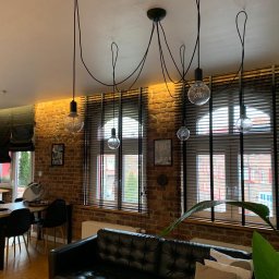 Igor Kravets - Loftowe wnętrze z ceglaną ścianą i oknami przesłoniętymi ciemnymi żaluzjami drewnianymi. Nad oknami zwisają lampy z dekoracyjnymi żarówkami.