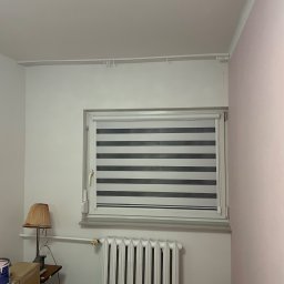 Igor Kravets - Pokój z oknem i zamontowaną roletą dzień-noc w szaro-białe pasy. Widoczny grzejnik, lampka nocna i puszka z farbą. Ściany w odcieniach bieli i delikatnego różu.