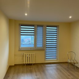 Igor Kravets - Jasne wnętrze z oknami przesłoniętymi roletami dzień-noc. Widoczny grzejnik pod oknem i krzesło z boku. Parkiet na podłodze. Świeże, żółte ściany i punktowe oświetlenie sufitowe.