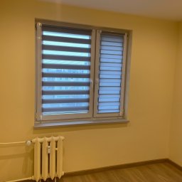 Igor Kravets - Zamontowane rolety dzień-noc w oknie, widoczne poziome pasy tkaniny w odcieniach szarości. Żółte ściany i drewniana podłoga. Grzejnik pod oknem.