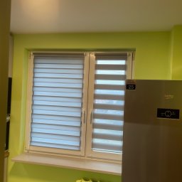 Igor Kravets - Zamontowane rolety dzień noc w oknie, widoczne z bliska w Katowicach. Na ścianie zielony kolor, obok stoi lodówka. Grzejnik pod oknem.