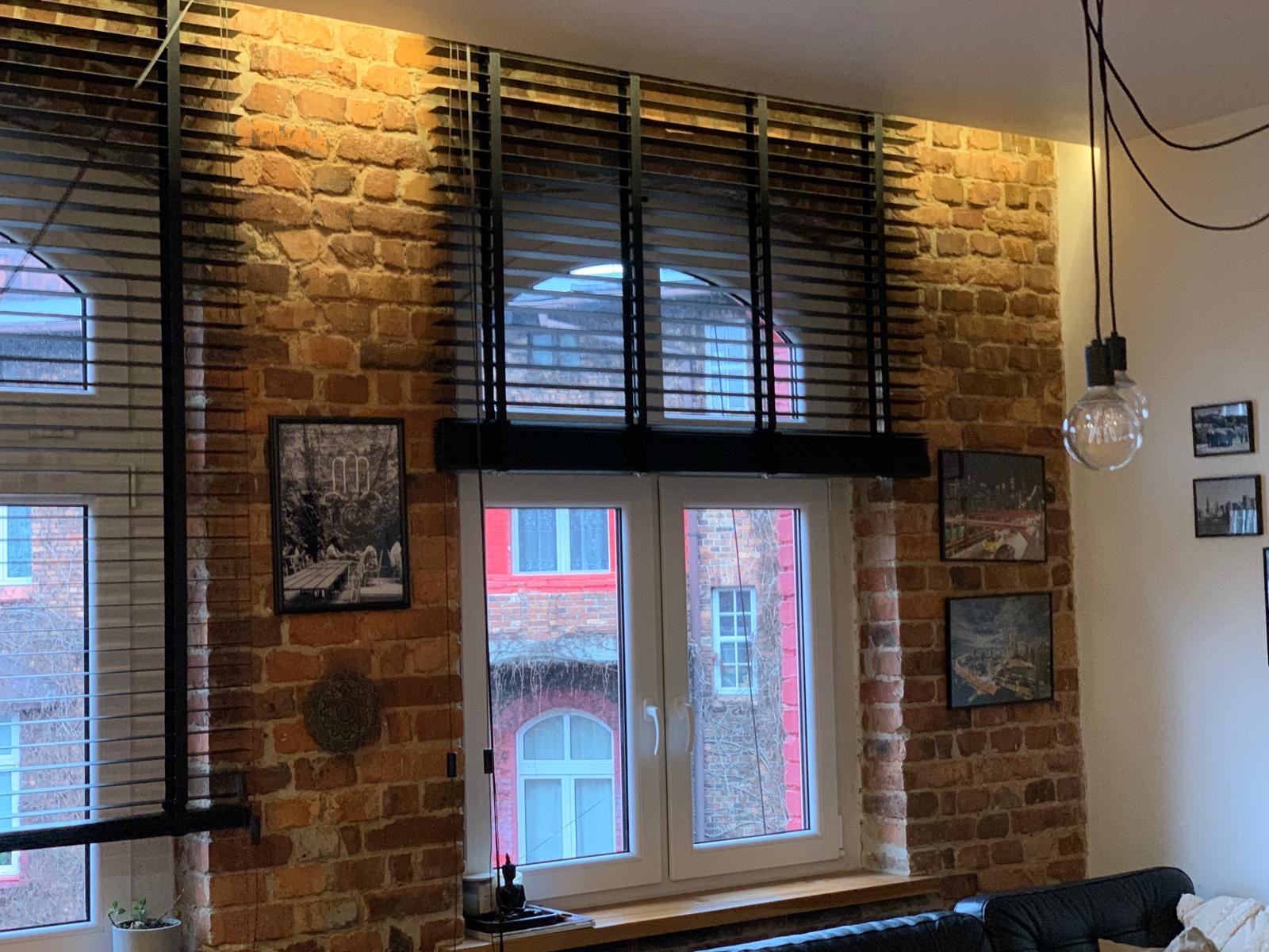 Ciemne żaluzje poziome zamontowane w oknach na tle ceglanej ściany w loftowym wnętrzu. Widok na budynki z czerwonej cegły za oknem.