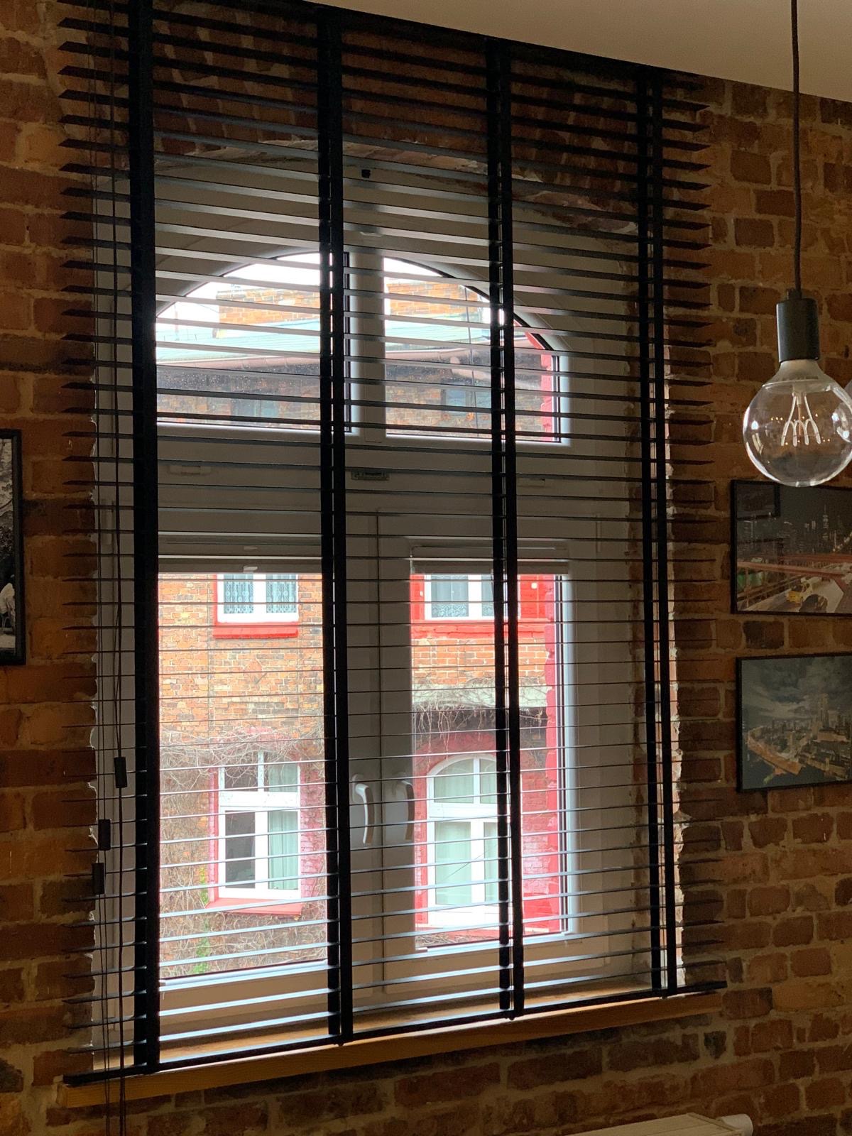 Ciemne żaluzje drewniane na tle ceglanej ściany i okna z widokiem na budynek. Lampa wisząca z żarówką Edisona po prawej stronie kadru.