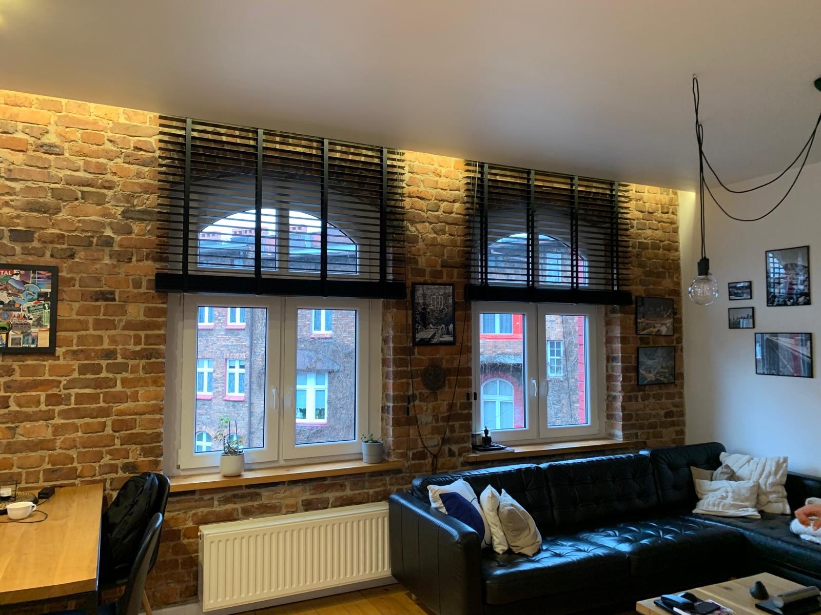 Czarne żaluzje drewniane w oknach o łukowym kształcie, na tle ceglanej ściany w salonie. Widok na budynek z zewnątrz. Ciepłe oświetlenie podkreśla fakturę cegły.