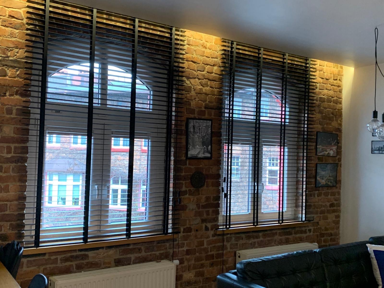 Dwa okna z czarnymi żaluzjami drewnianymi na tle ściany z czerwonej cegły. Widok na budynki za oknem. Ciepłe oświetlenie podkreśla fakturę cegły.