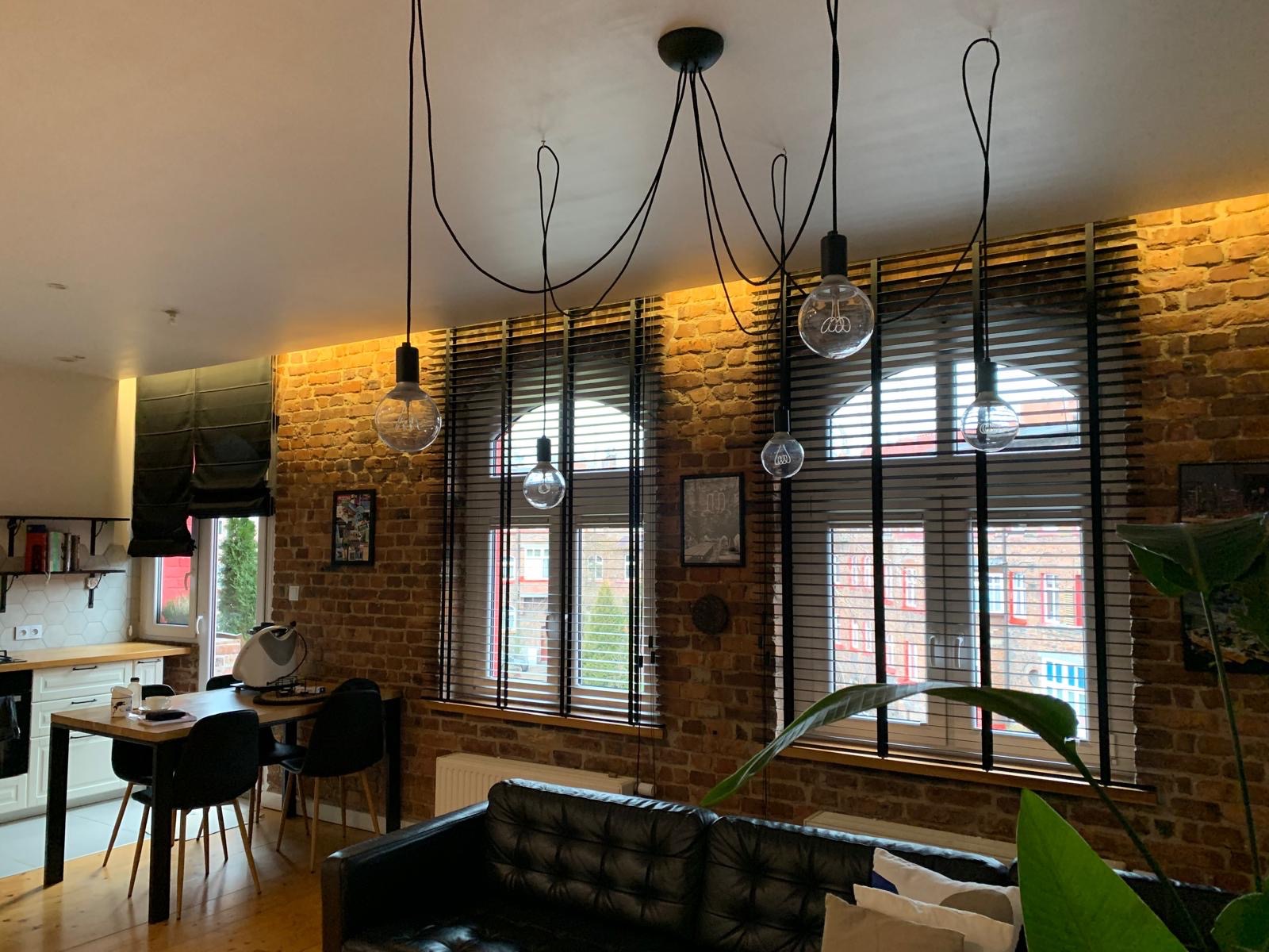 Loftowe wnętrze z ceglaną ścianą i oknami przesłoniętymi ciemnymi żaluzjami drewnianymi. Nad oknami zwisają lampy z dekoracyjnymi żarówkami.