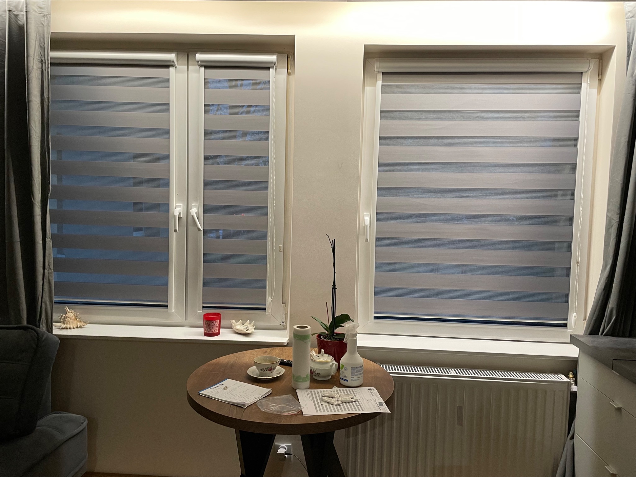 Dwa okna z zamontowanymi roletami dzień-noc w odcieniach szarości. Na parapecie i stole dekoracje, w tle zasłony. Widok na wnętrze mieszkania.