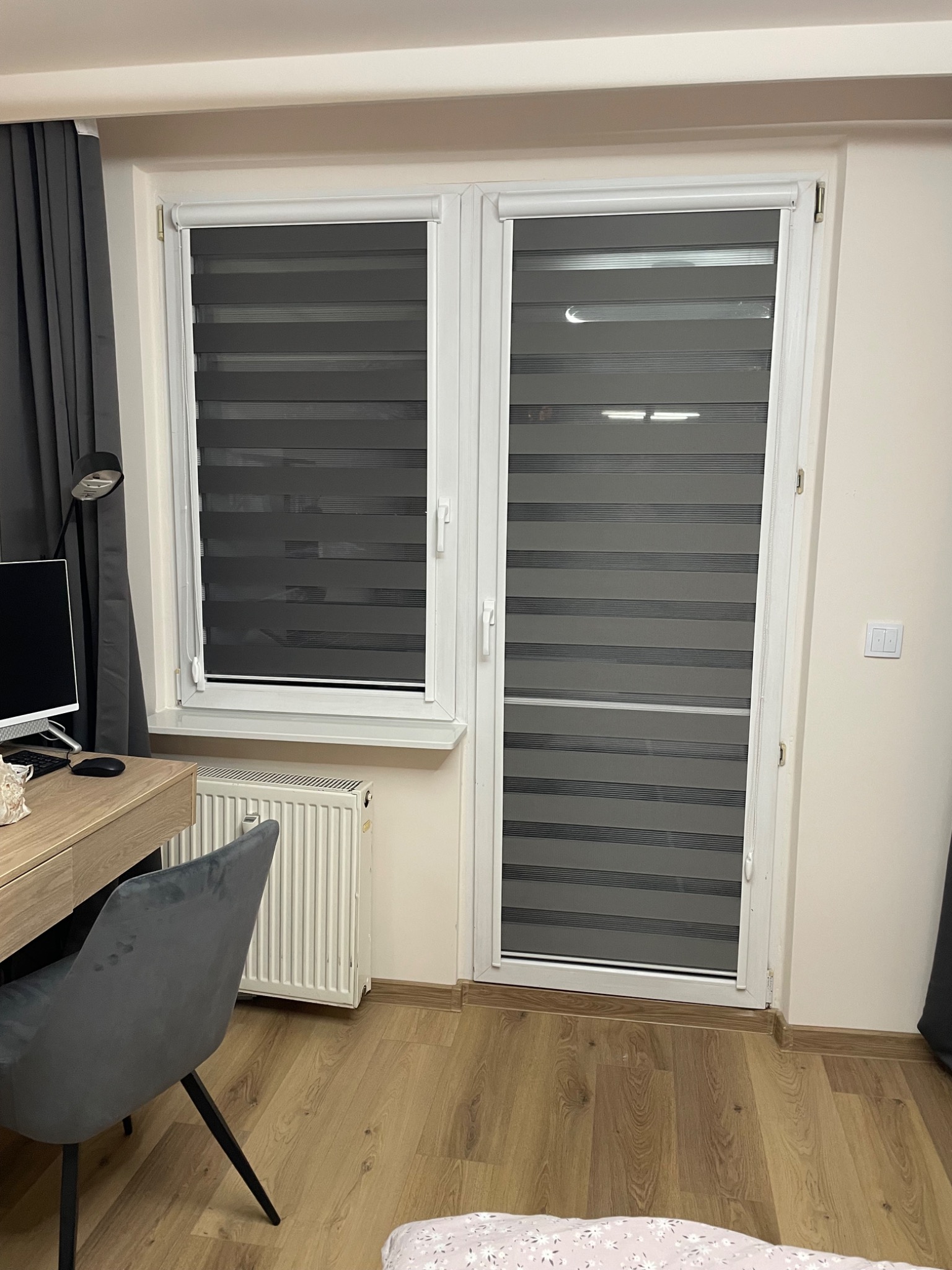 Zamknięte rolety dzień-noc w oknie PCV, widoczne poziome pasy w odcieniach szarości, biurko z komputerem i krzesło w tle, wnętrze pokoju.