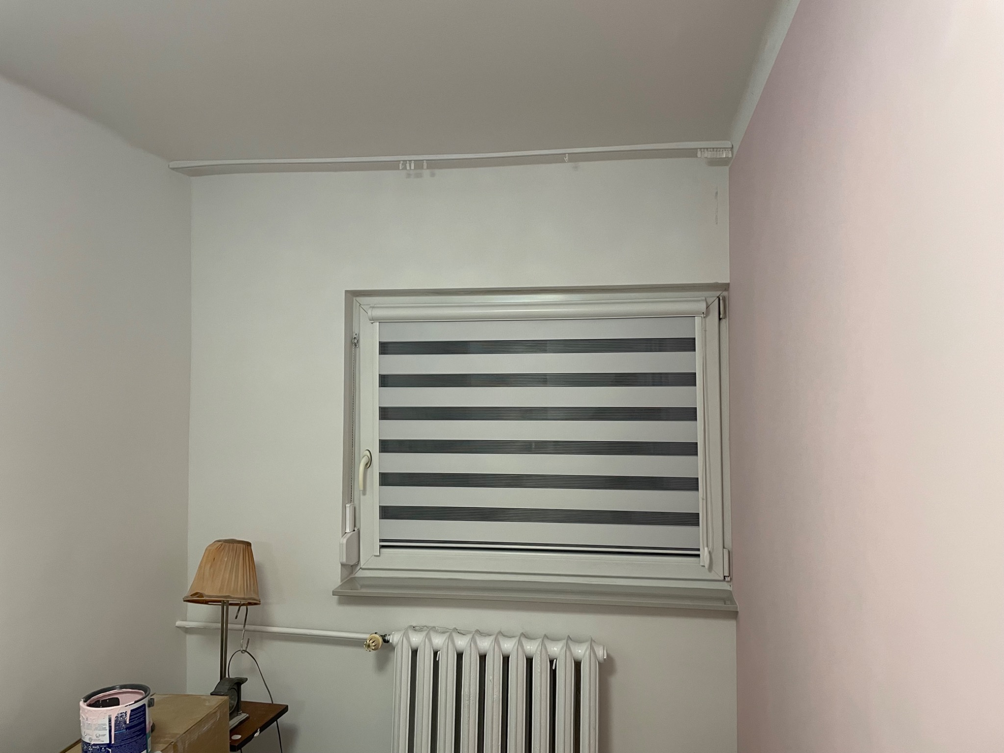 Pokój z oknem i zamontowaną roletą dzień-noc w szaro-białe pasy. Widoczny grzejnik, lampka nocna i puszka z farbą. Ściany w odcieniach bieli i delikatnego różu.