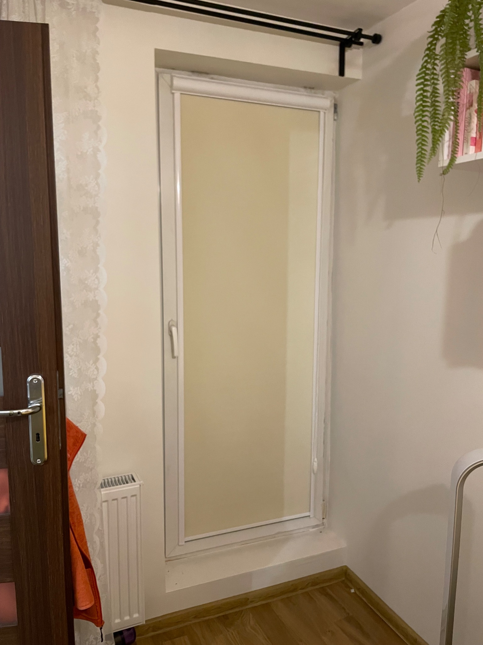 Zamknięta, wewnętrzna roleta w białej ramie, zamontowana w pokoju z drewnianą podłogą i grzejnikiem. Roleta w kolorze kremowym, zasłania okno.