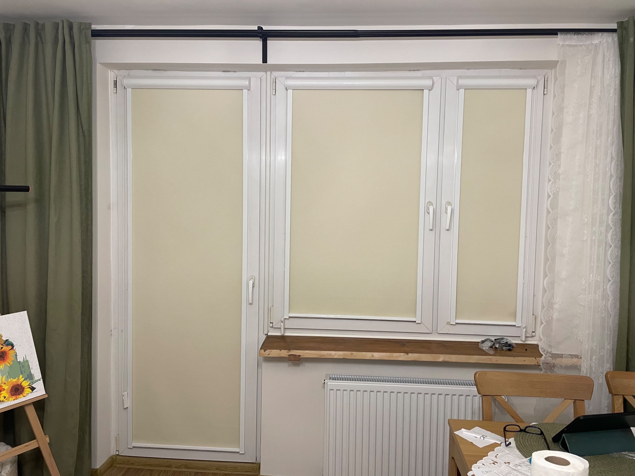 Beżowe rolety wewnętrzne w białych ramach okiennych, widok z bliska. Po lewej stronie zasłona w odcieniach zieleni, po prawej firanka. Na parapecie leżą drobne przedmioty.