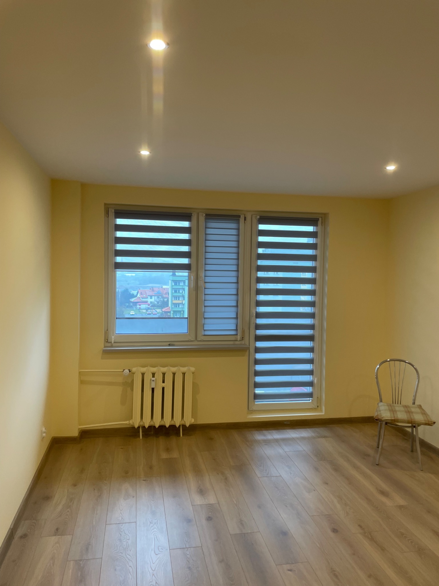 Jasne wnętrze z oknami przesłoniętymi roletami dzień-noc. Widoczny grzejnik pod oknem i krzesło z boku. Parkiet na podłodze. Świeże, żółte ściany i punktowe oświetlenie sufitowe.