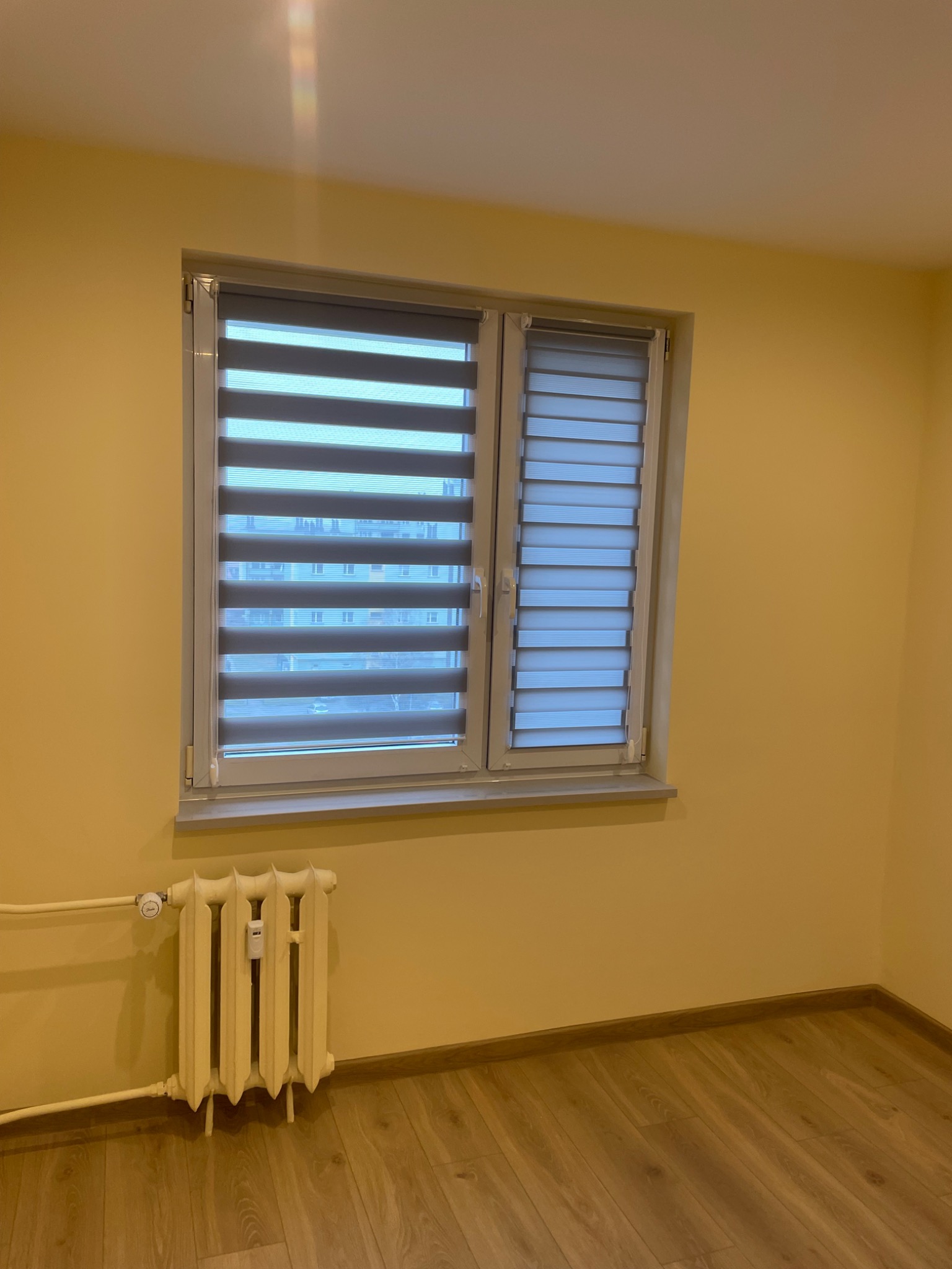 Zamontowane rolety dzień-noc w oknie, widoczne poziome pasy tkaniny w odcieniach szarości. Żółte ściany i drewniana podłoga. Grzejnik pod oknem.