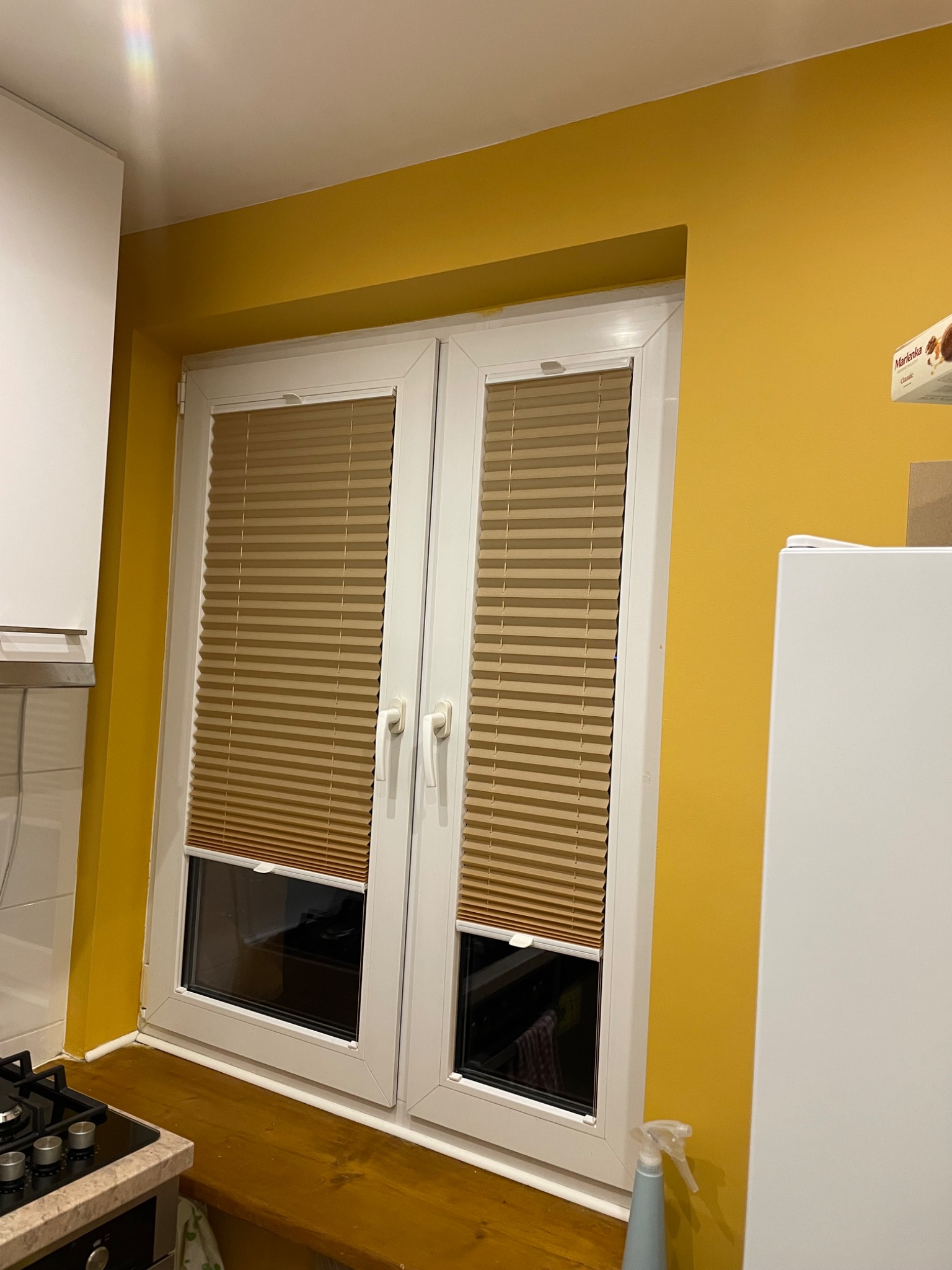 Beżowe plisy okienne zamontowane w białych oknach na tle żółtej ściany w kuchni. Widoczny fragment blatu i kuchenki. Ujęcie z bliska, z widocznymi detalami plis.
